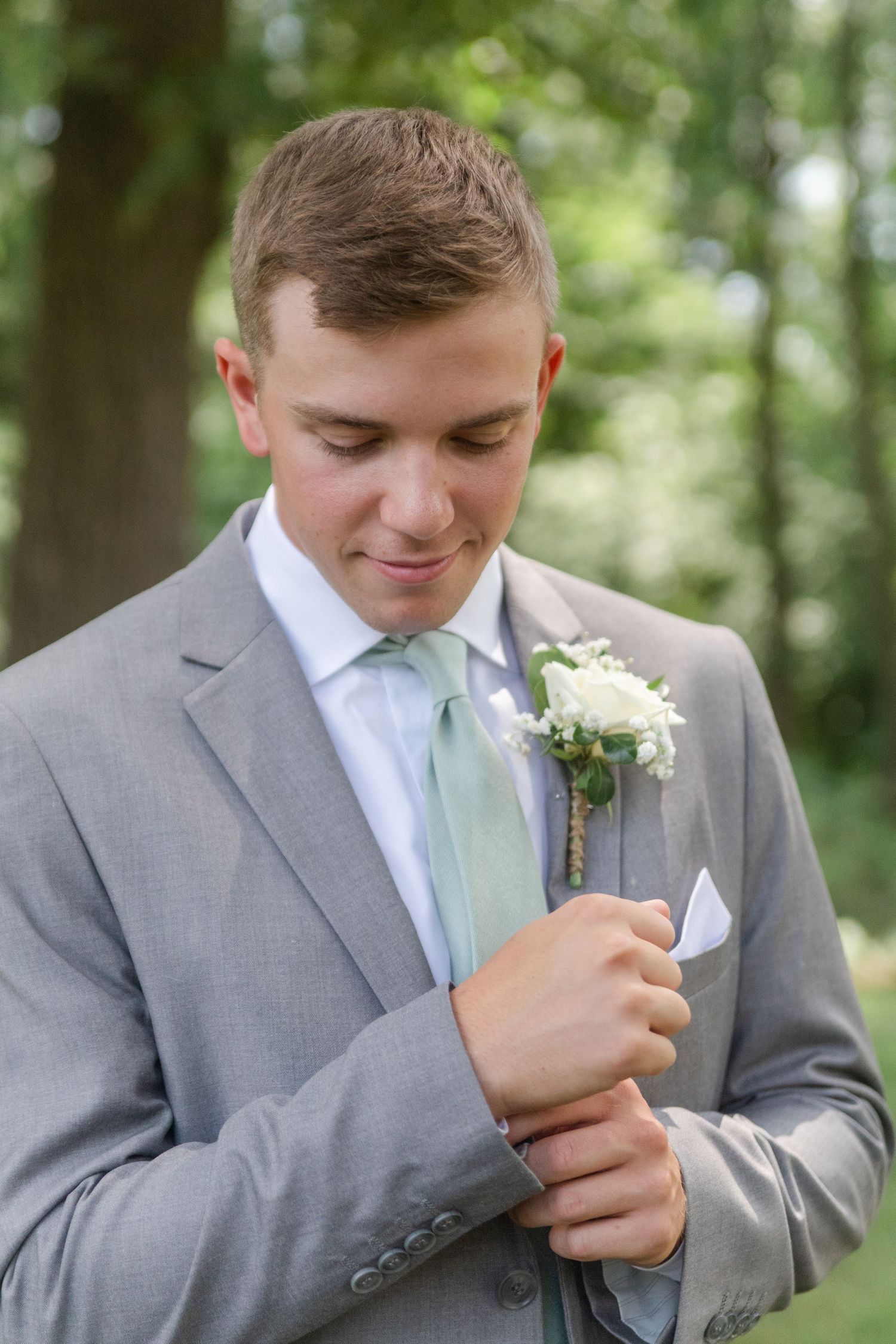 A groomsman in a gray suit and mint tie pins a white boutonniere to his lapel.