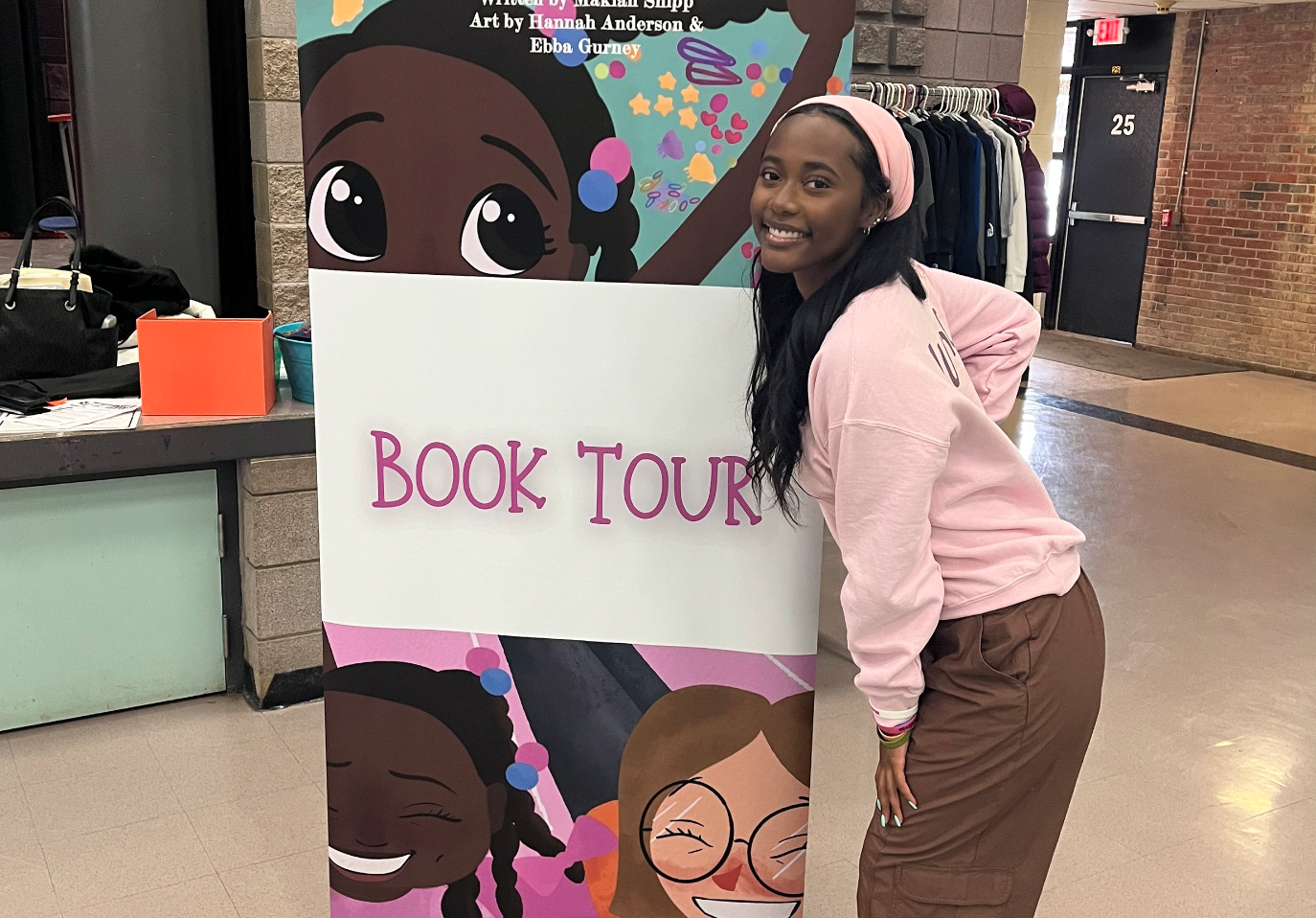 An author stands next to a promotional book tour sign with decorative artwork.