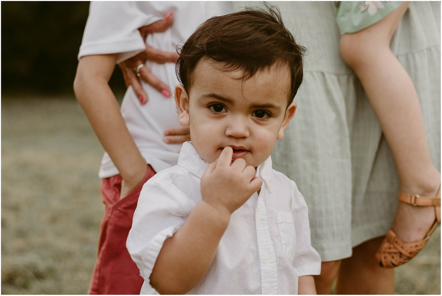 A close-up shot of a young child in a white shirt with hand near their mouth during a family photoshoot.