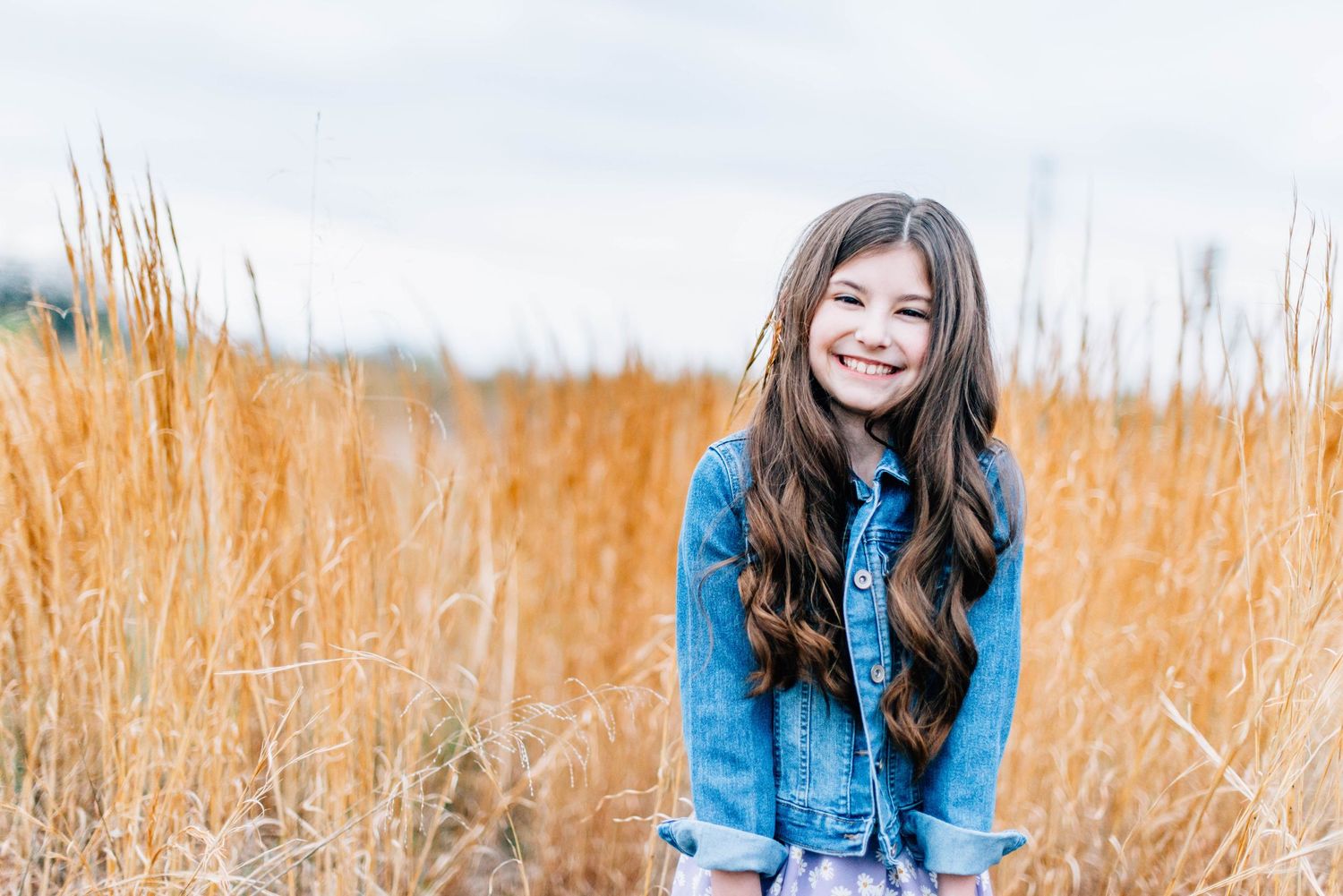 A young person stands in a golden wheat field wearing a denim jacket on a bright day.