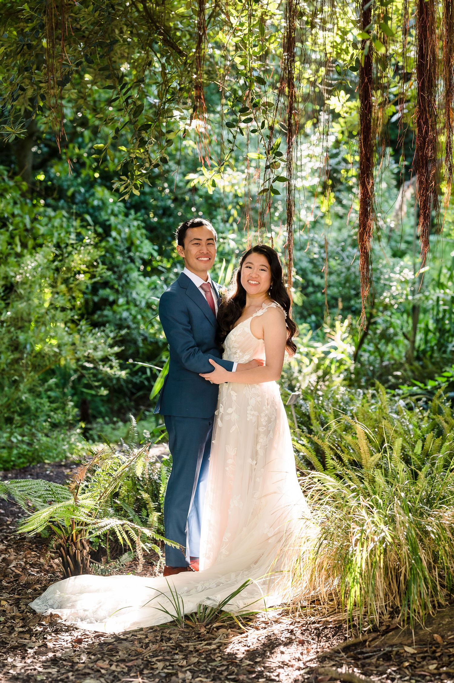 A wedding couple poses in magical forest lighting surrounded by ferns and trees.