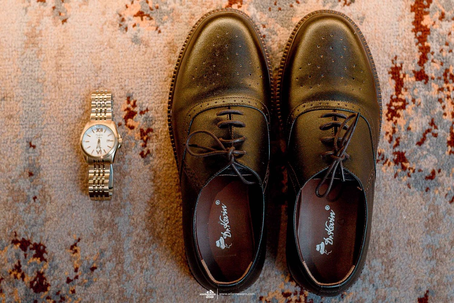 Brown leather dress shoes and gold watch arranged on a rustic pink surface.