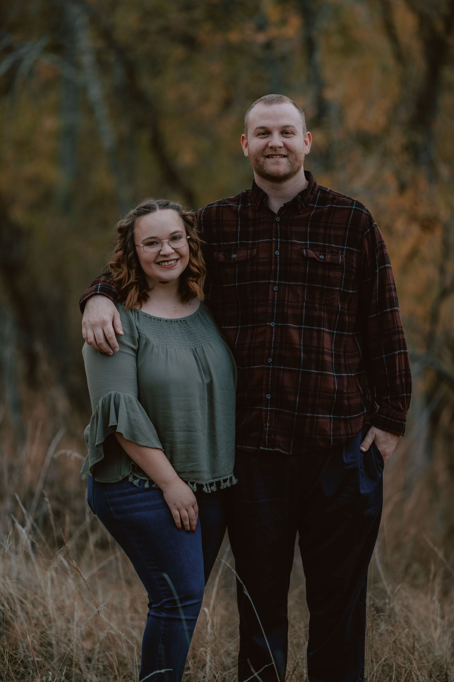 Smiling couple poses together in autumn field with colorful fall foliage in background.