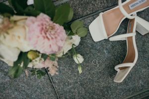 Wedding details including pink roses and white strappy bridal shoes on a stone surface.