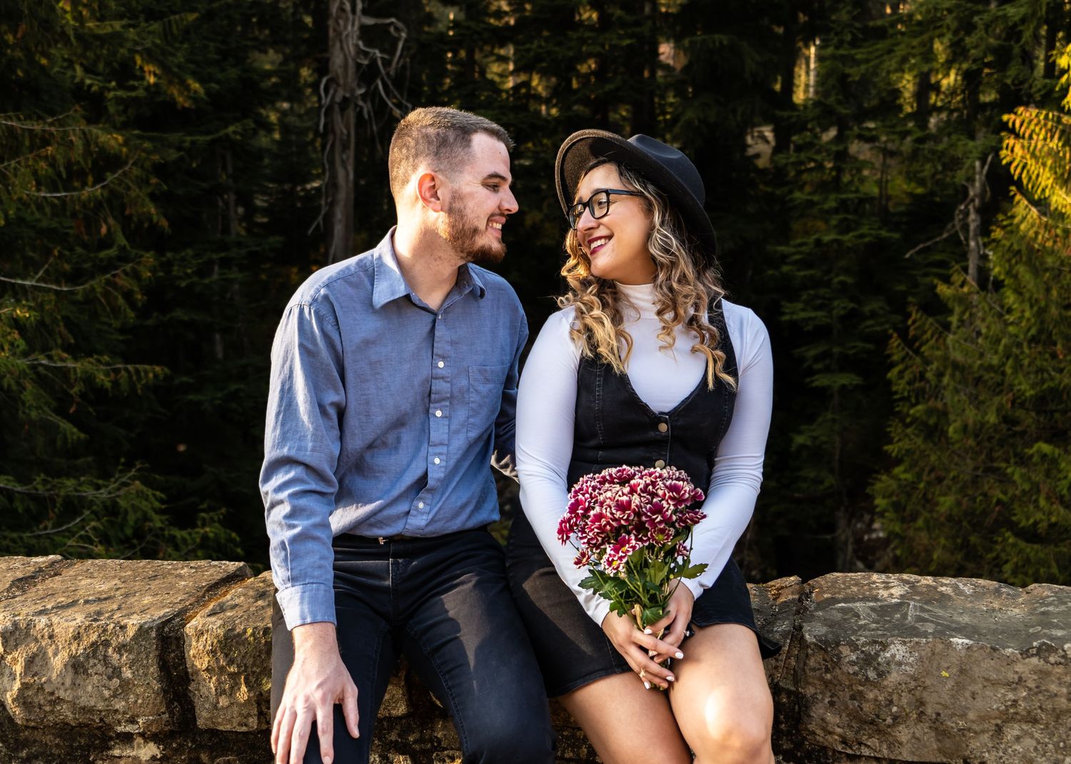 Couple sitting on stone wall with pink flowers surrounded by evergreen forest.