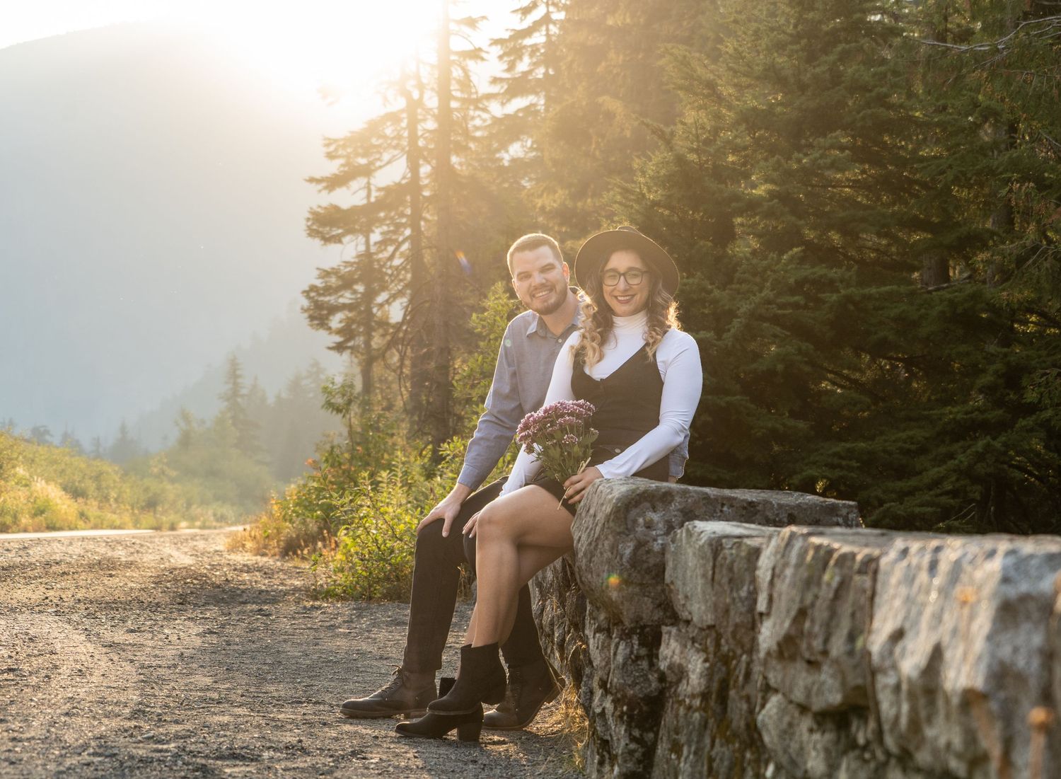Couple sitting on stone wall during golden hour with evergreen forest backdrop.