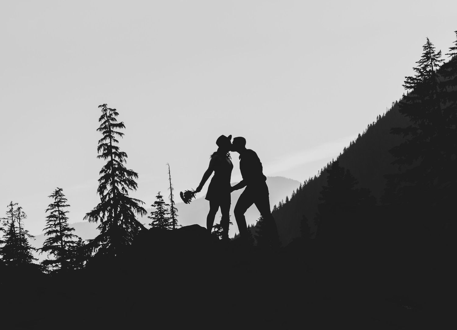 Silhouetted couple standing on hilltop against mountain landscape at dusk.