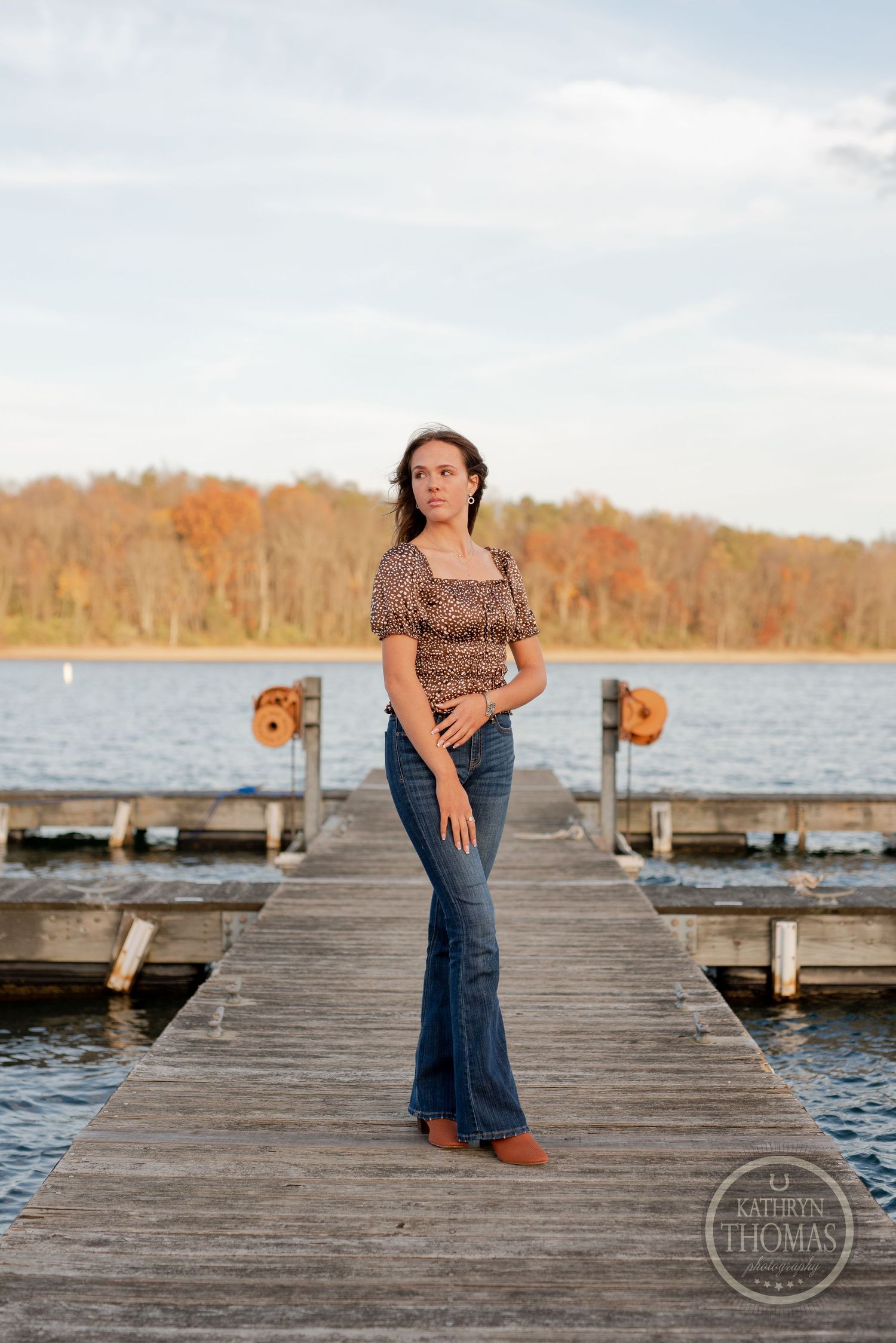 Full length portrait on a wooden pier with water and autumn trees in the background.