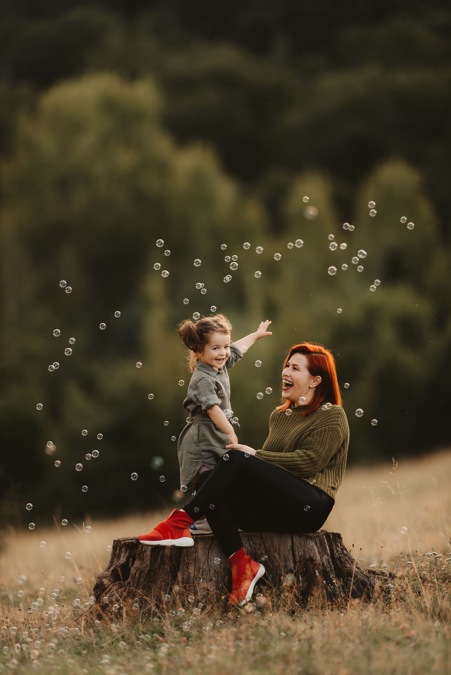 A parent and child play with bubbles while sitting on a tree stump outdoors.