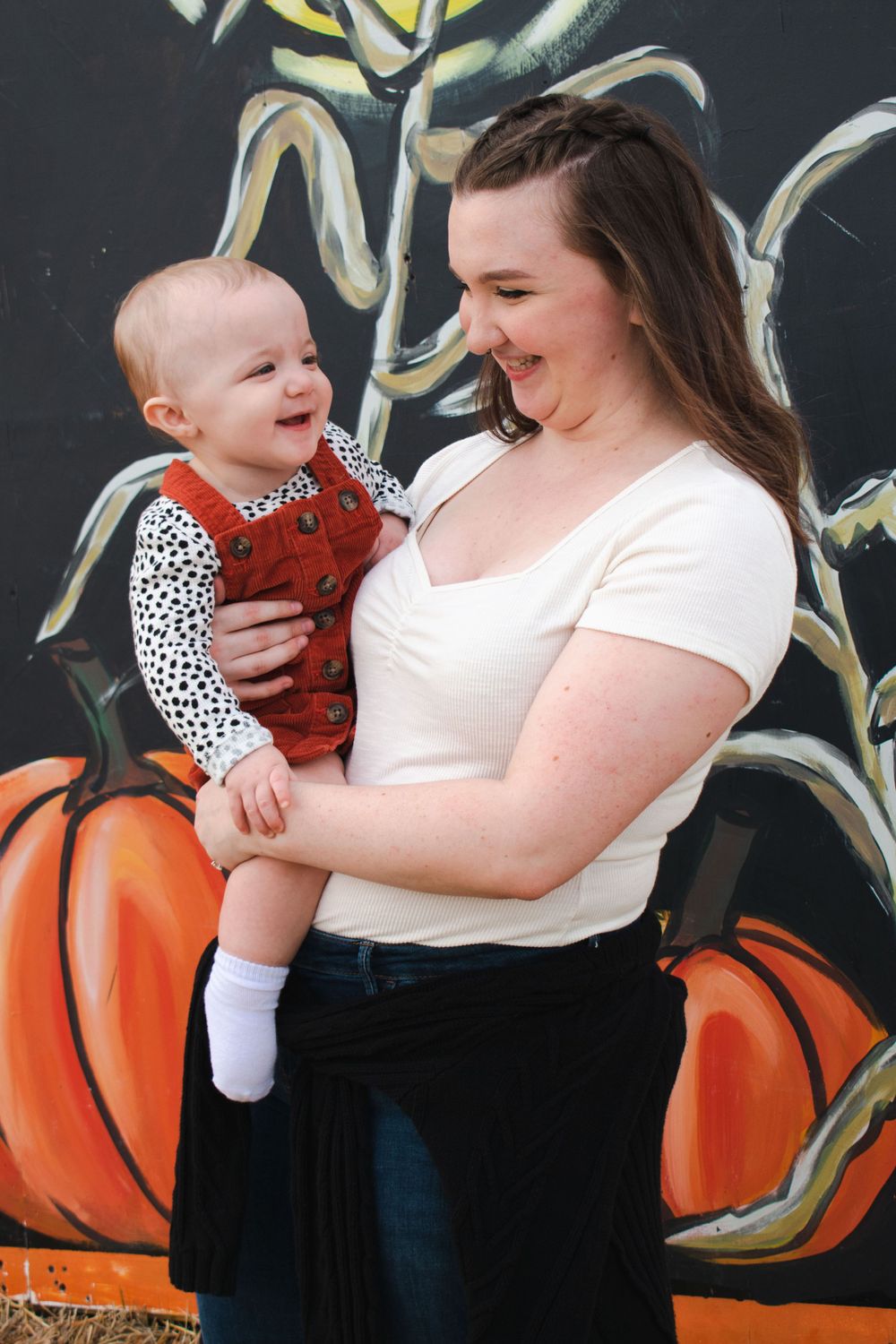 A young mom giggles along with her baby in front of a pumpkin mural