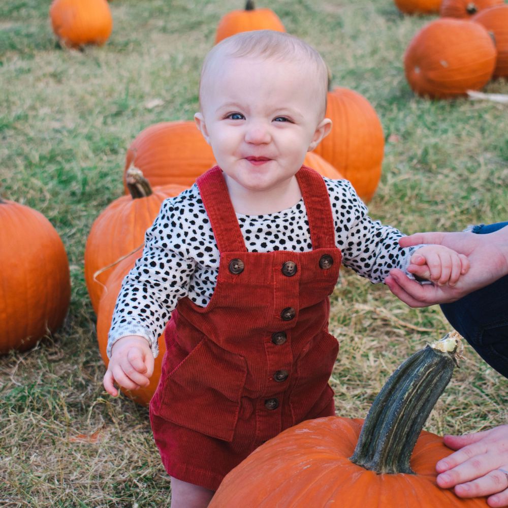 A cheeky baby girl grins for camera, surrounded by pumpkins