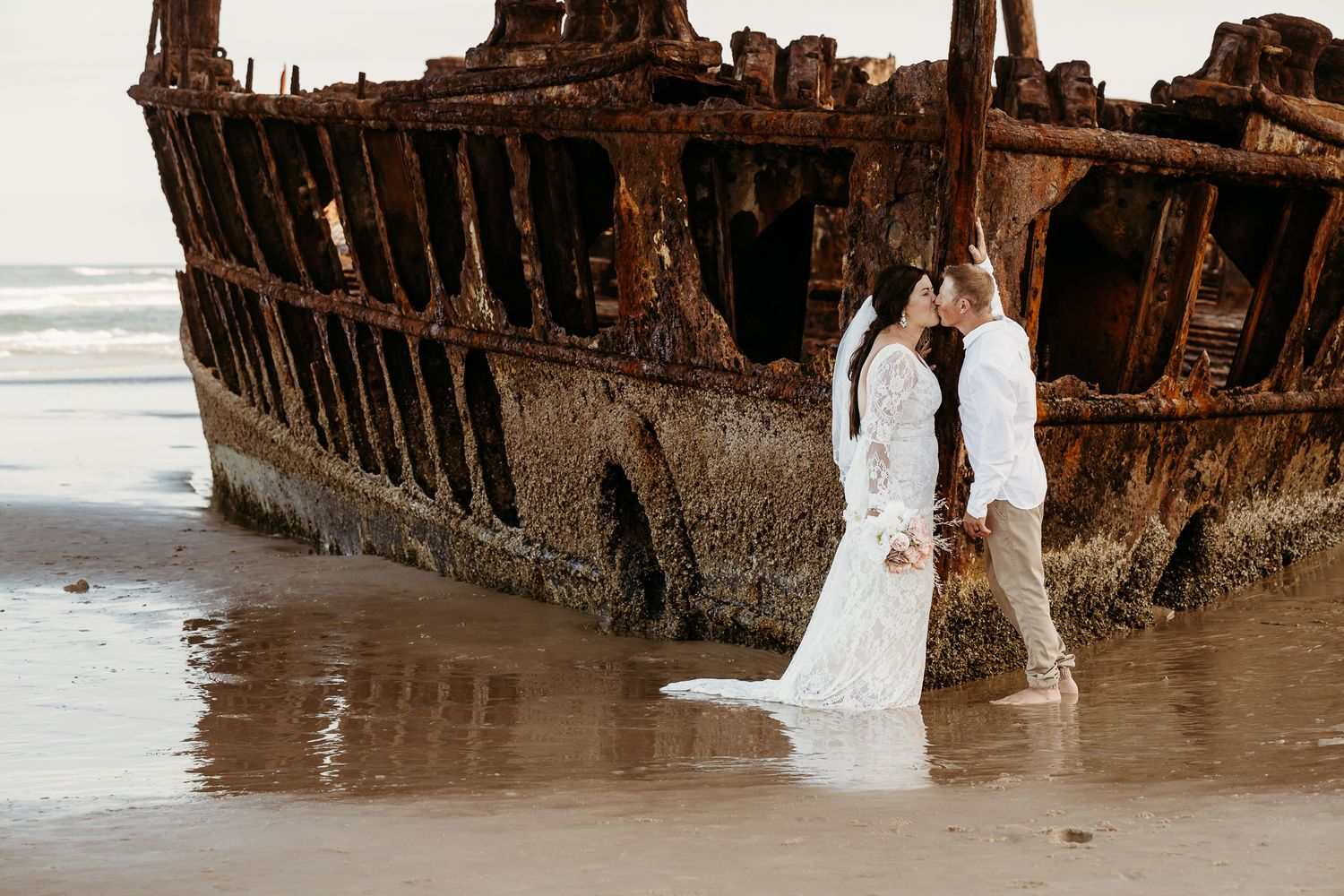 Wedding couple poses by rustic shipwreck on beach at sunset.