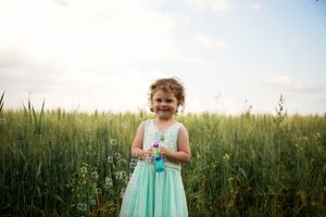 Flower girl stands laughing in front of a wheat field. She hold a bubble gun, bubbles fly all around her.