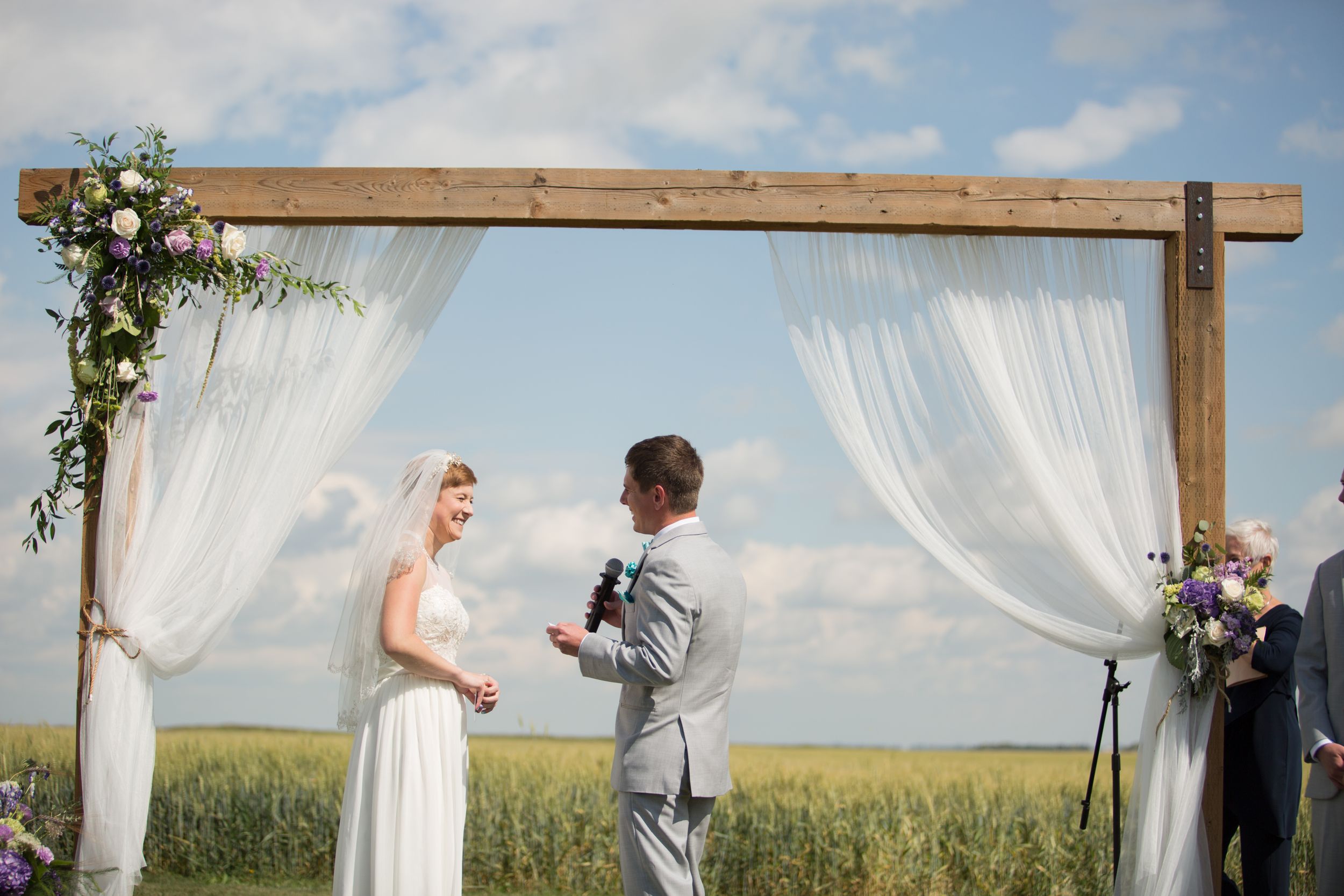 Bride and groom stand facing each other in front of a wheat field under a wedding arch. Groom holds a microphone and is speaking his vows.