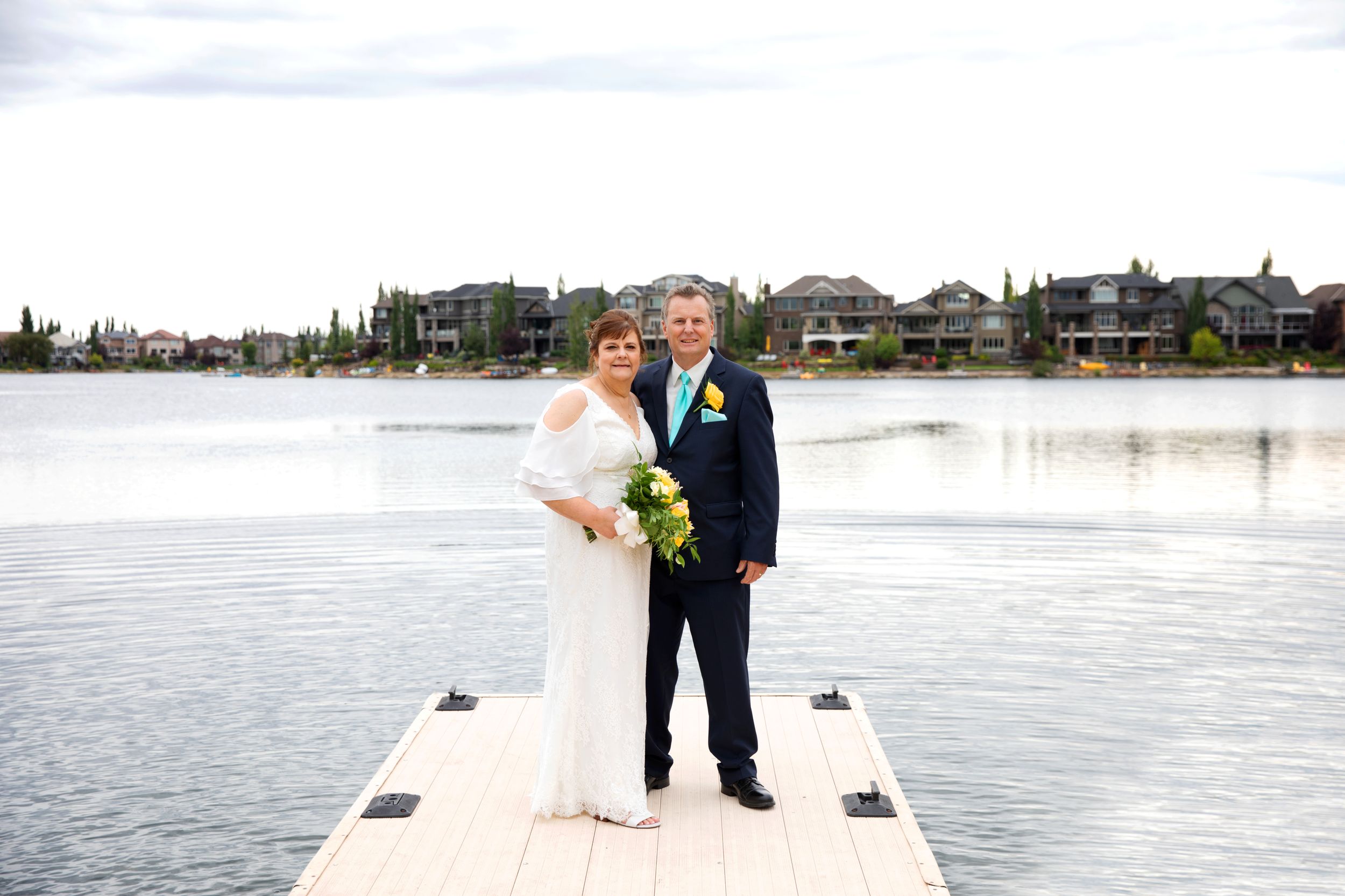Bride and groom stand together at the end of a floating dock . Behind them is a beautiful lake on a sunny day.