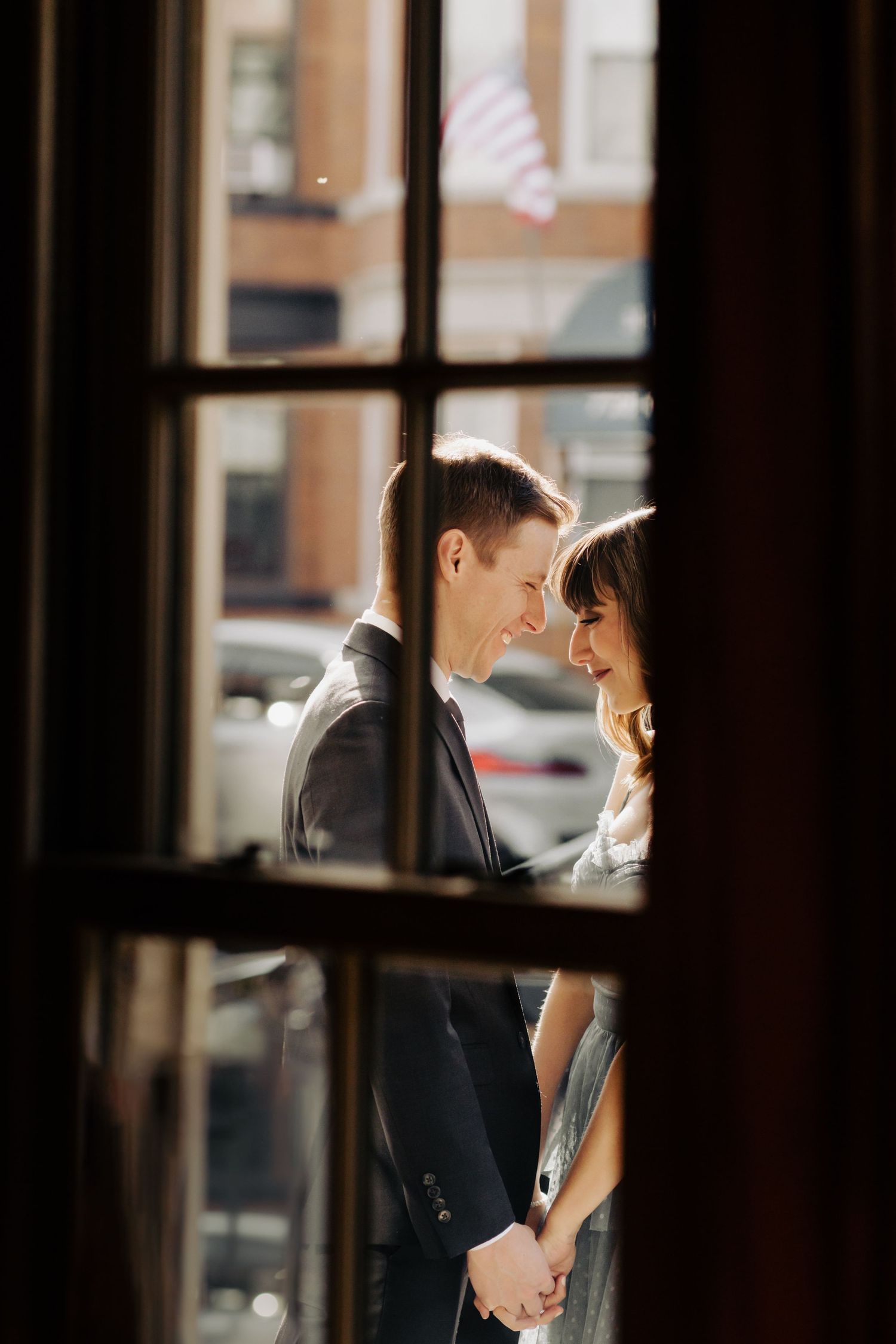 Couple in formal attire sharing a tender moment by large windows.