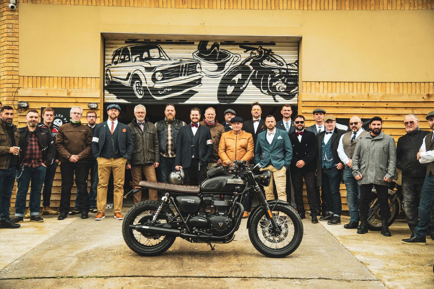 Motorcycle enthusiasts gathered around a custom black motorcycle against a vintage-themed wall mural.