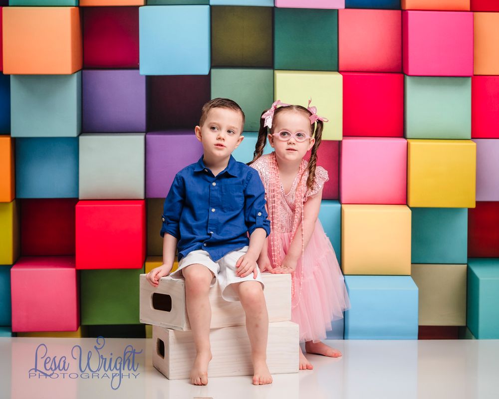 Two children seated on bench against colorful geometric block wall background.