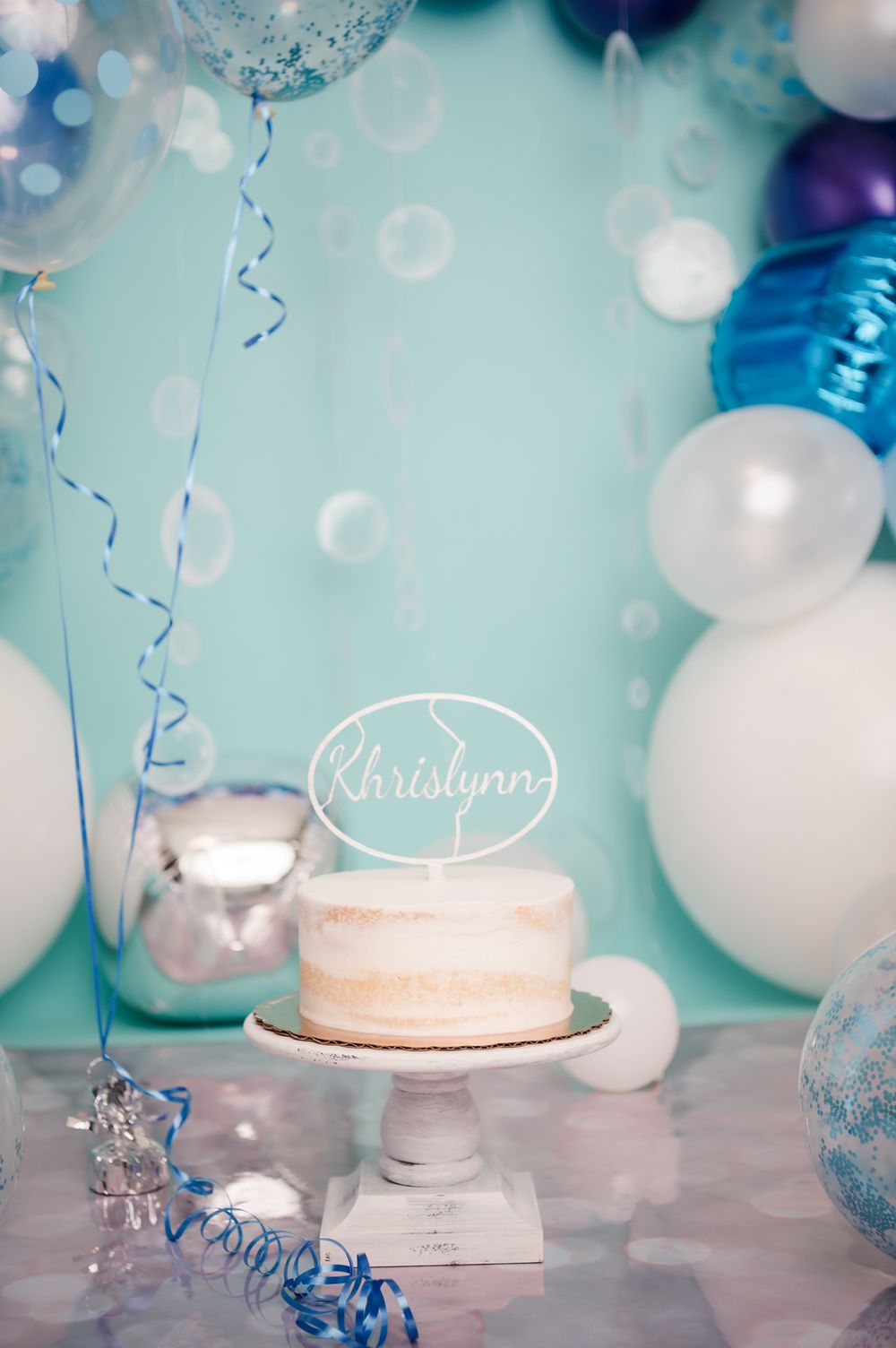 Elegant birthday cake with name topper surrounded by blue and white balloons on a pedestal.