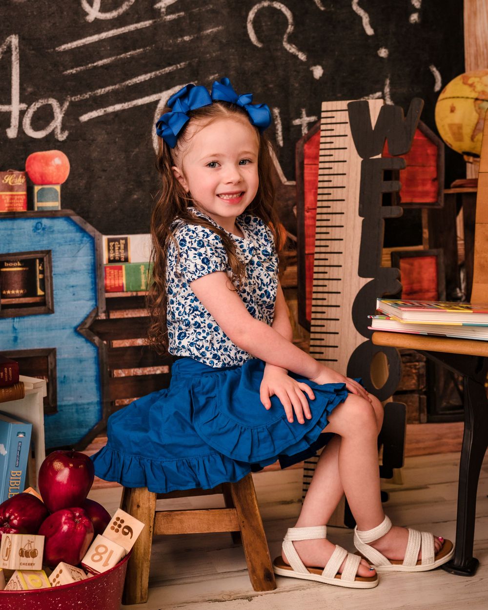 Student sits in a classroom setting with blue bow and dress against a chalkboard background with educational props.