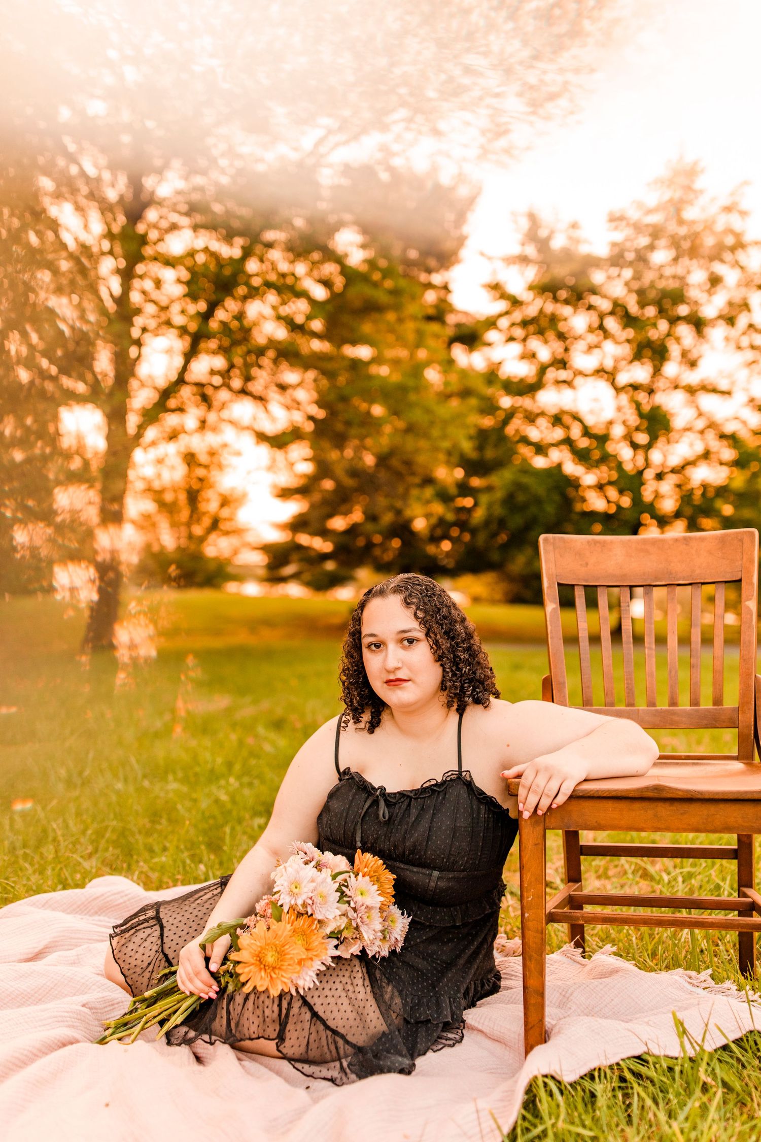 Young woman in black dress sits on blanket with flowers beside wooden chair at golden hour.