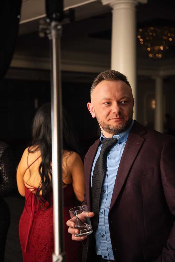 Person in burgundy suit holds drink while posing in dimly lit venue with columns.