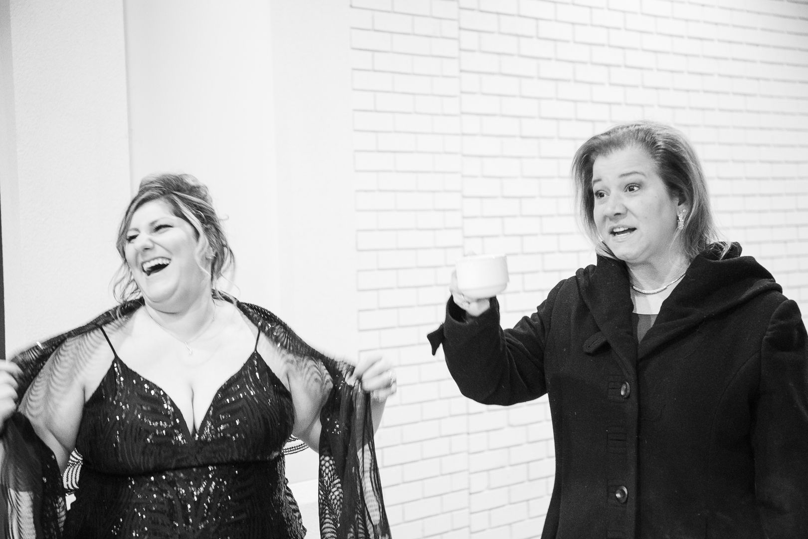 Two people share a candid laughing moment in black and white against a white brick wall.