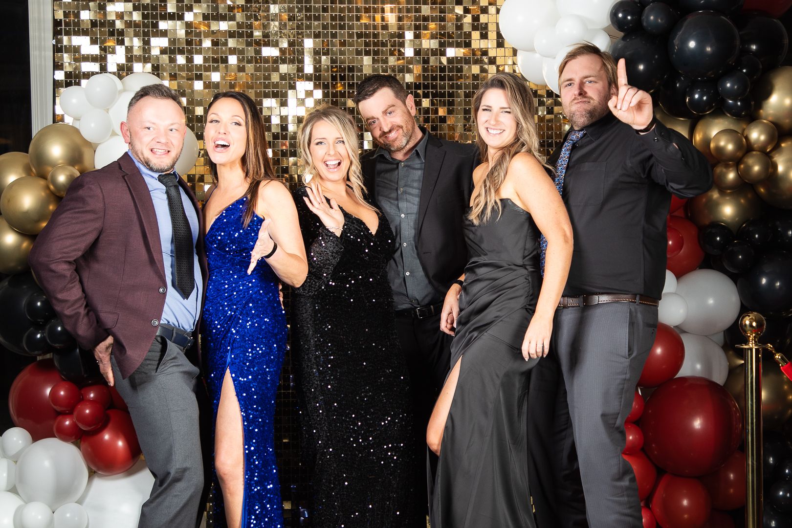 Group in formal attire poses against gold sequin wall with black and gold balloon decorations.