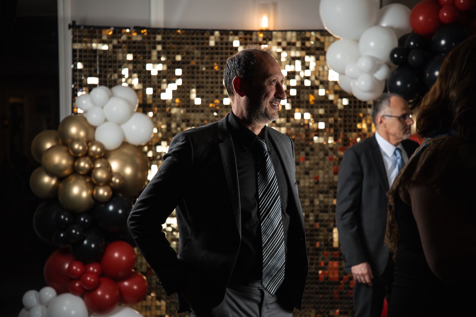 People gathered at event with gold sequin backdrop and balloon decorations.