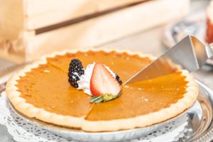 Pumpkin pie garnished with fresh berries and cream being cut on decorative silver platter.