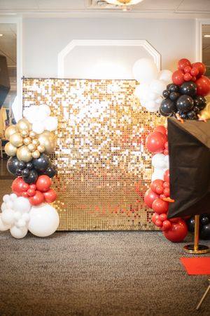 Event space with gold sequin backdrop and red, black, and gold balloon decorations.