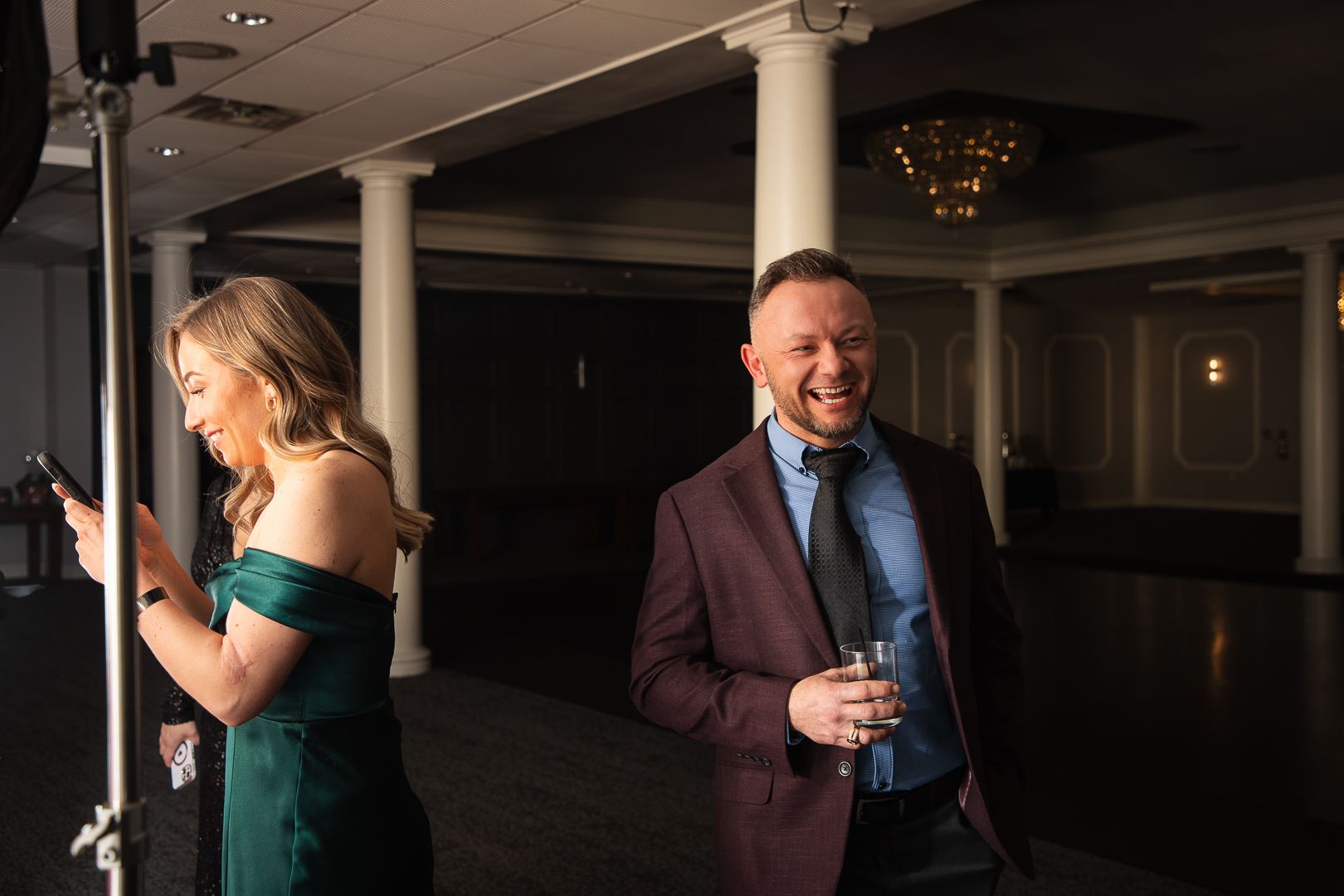 Two people enjoying drinks and laughing at formal event in venue with columns.