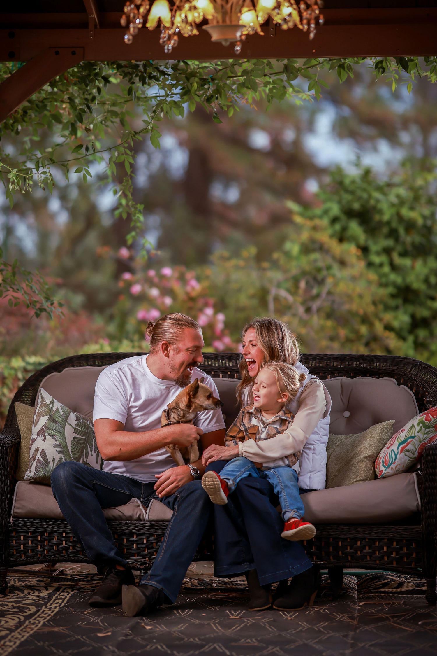 Happy family with a toddler and puppy sitting on a wicker sofa under a garden pergola.