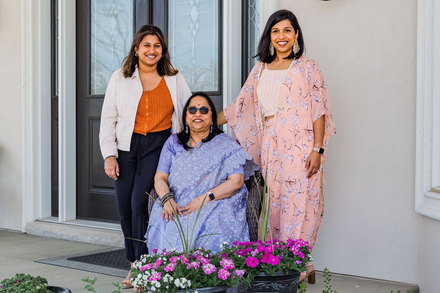 Three people posed together on front porch steps with colorful flowers and elegant doorway.