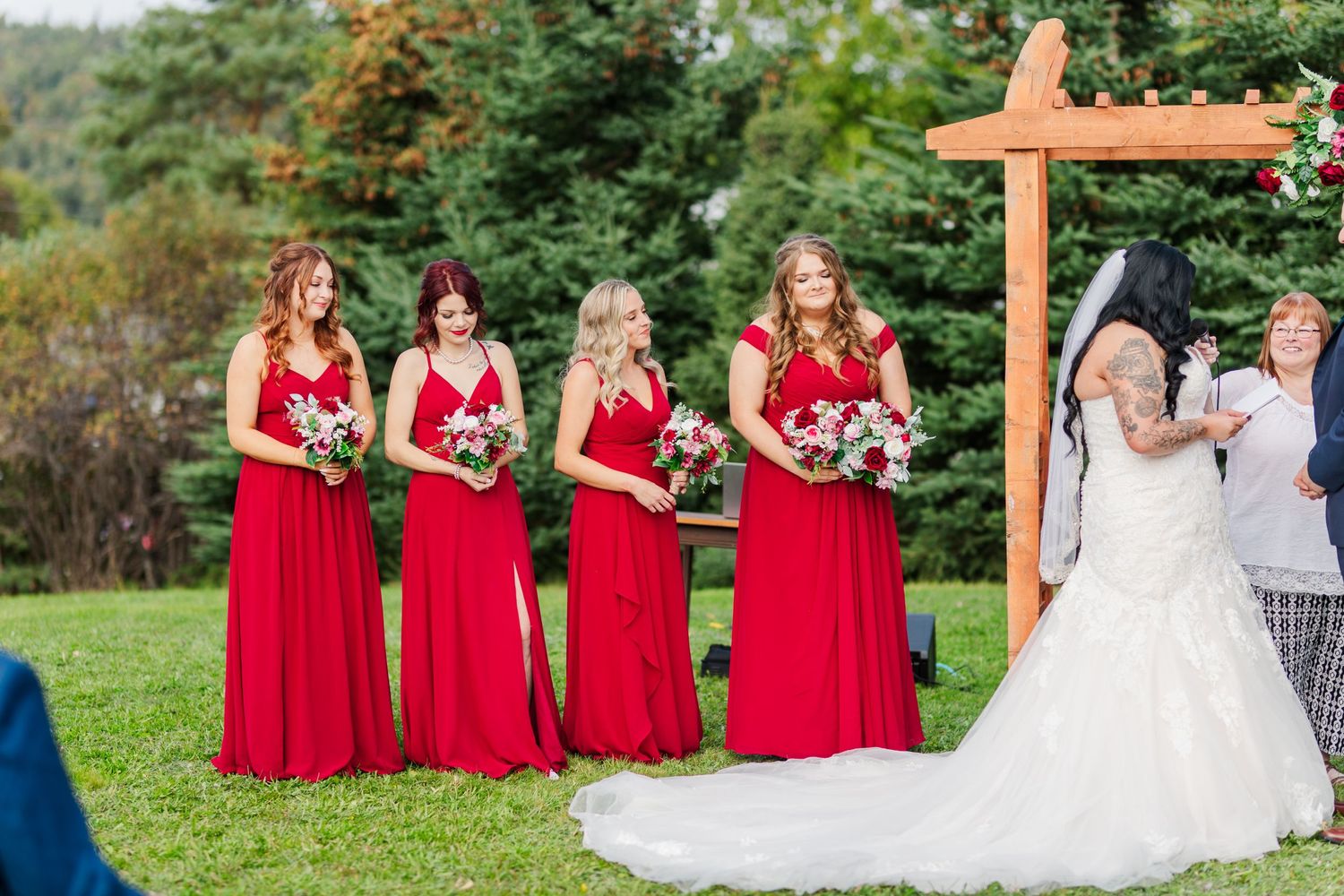 Bridesmaids in red dresses stand beside bride in white gown during outdoor ceremony.