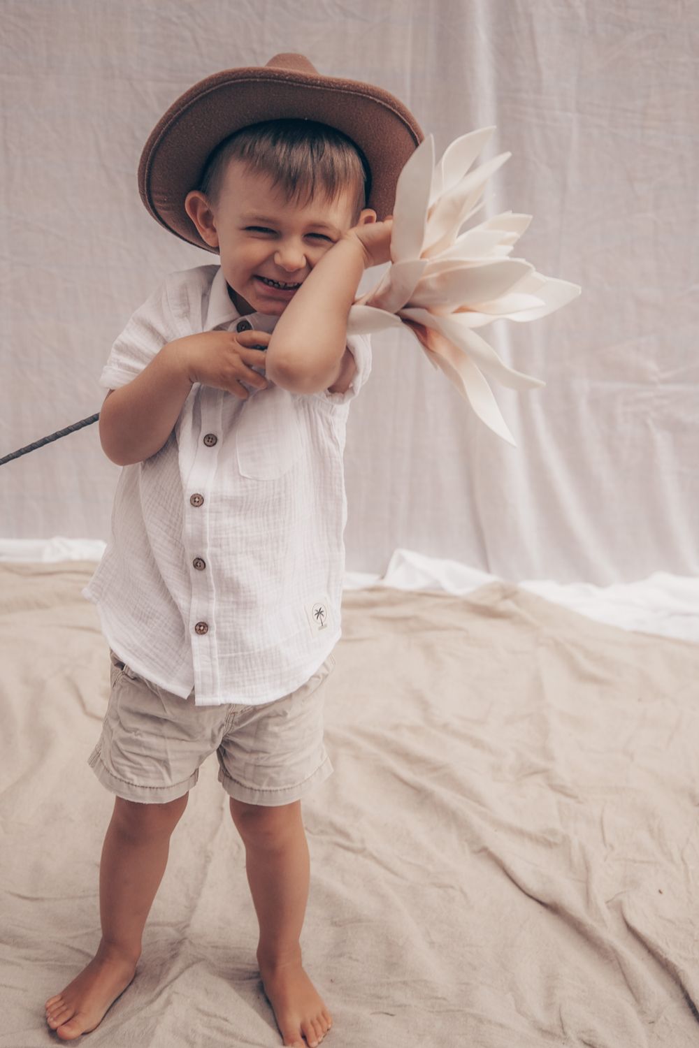 A joyful child wearing a brown hat and white shirt holds a large white flower while standing barefoot.