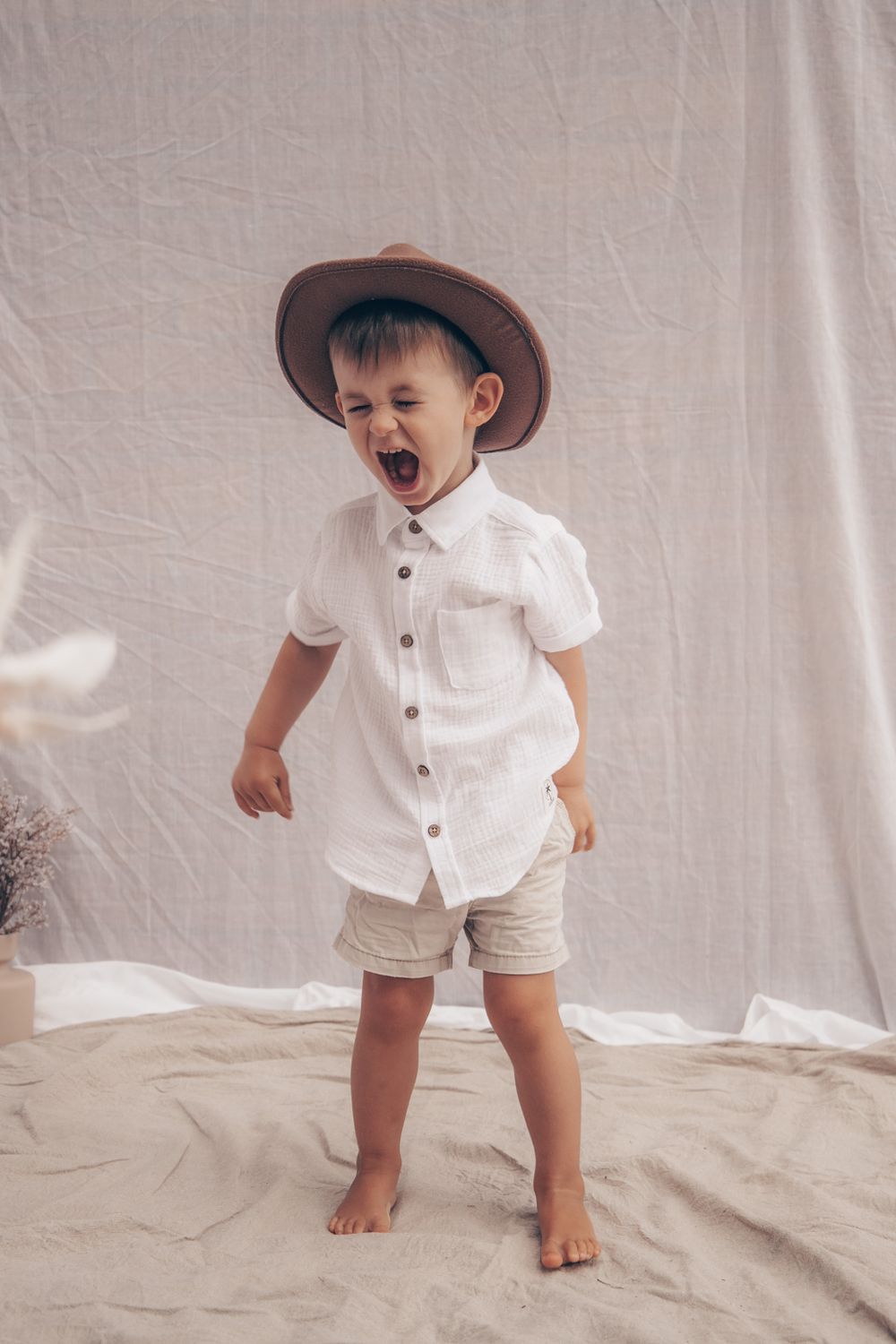 A laughing child in a brown hat and white outfit stands barefoot against a light backdrop.