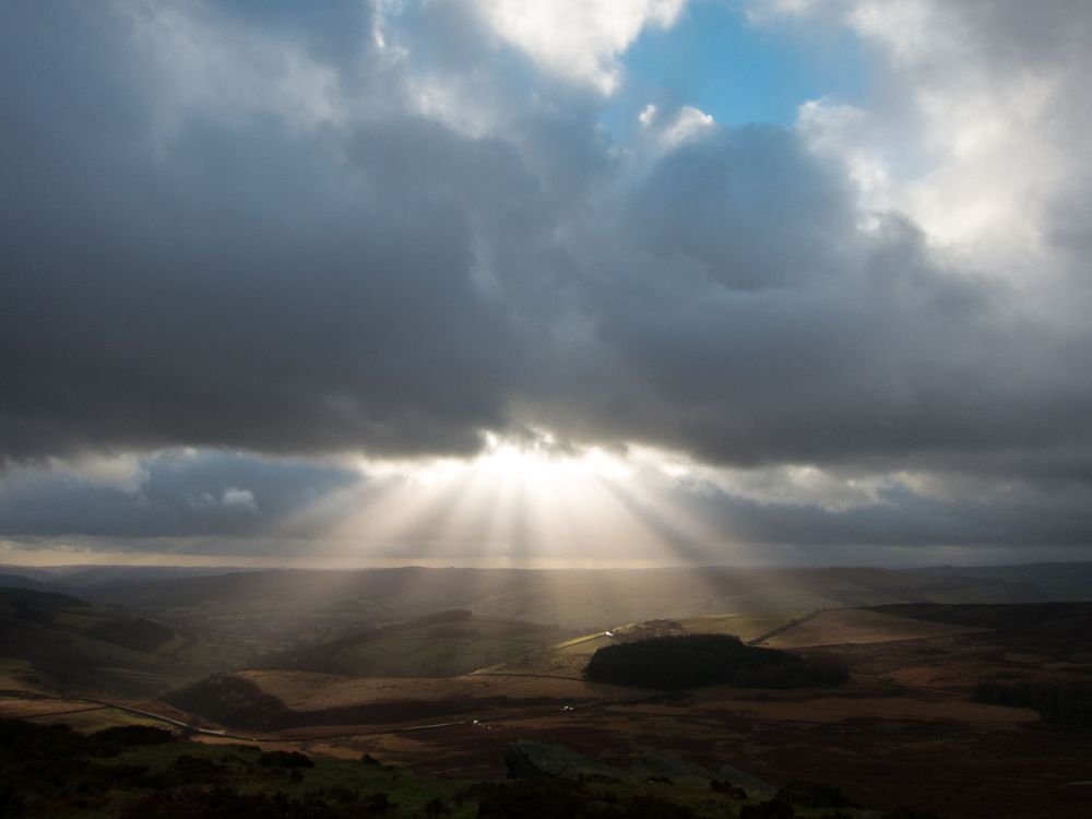 Dramatic sunbeams pierce through dark storm clouds over a remote moorland landscape in the Peak District.