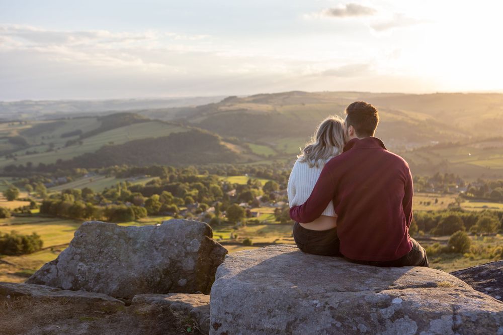 A couple sits together on a rocky overlook watching a scenic mountain valley sunset at Curbar Edge.