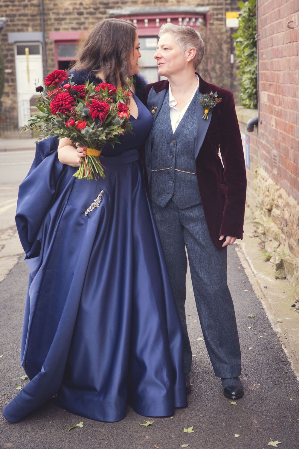 Two brides in formal attire sharing intimate moment against brick wall, with red bouquet.