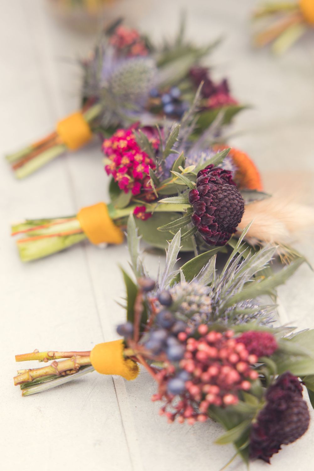 Colorful wedding boutonnieres with purple flowers, orange accents and greenery on white surface.