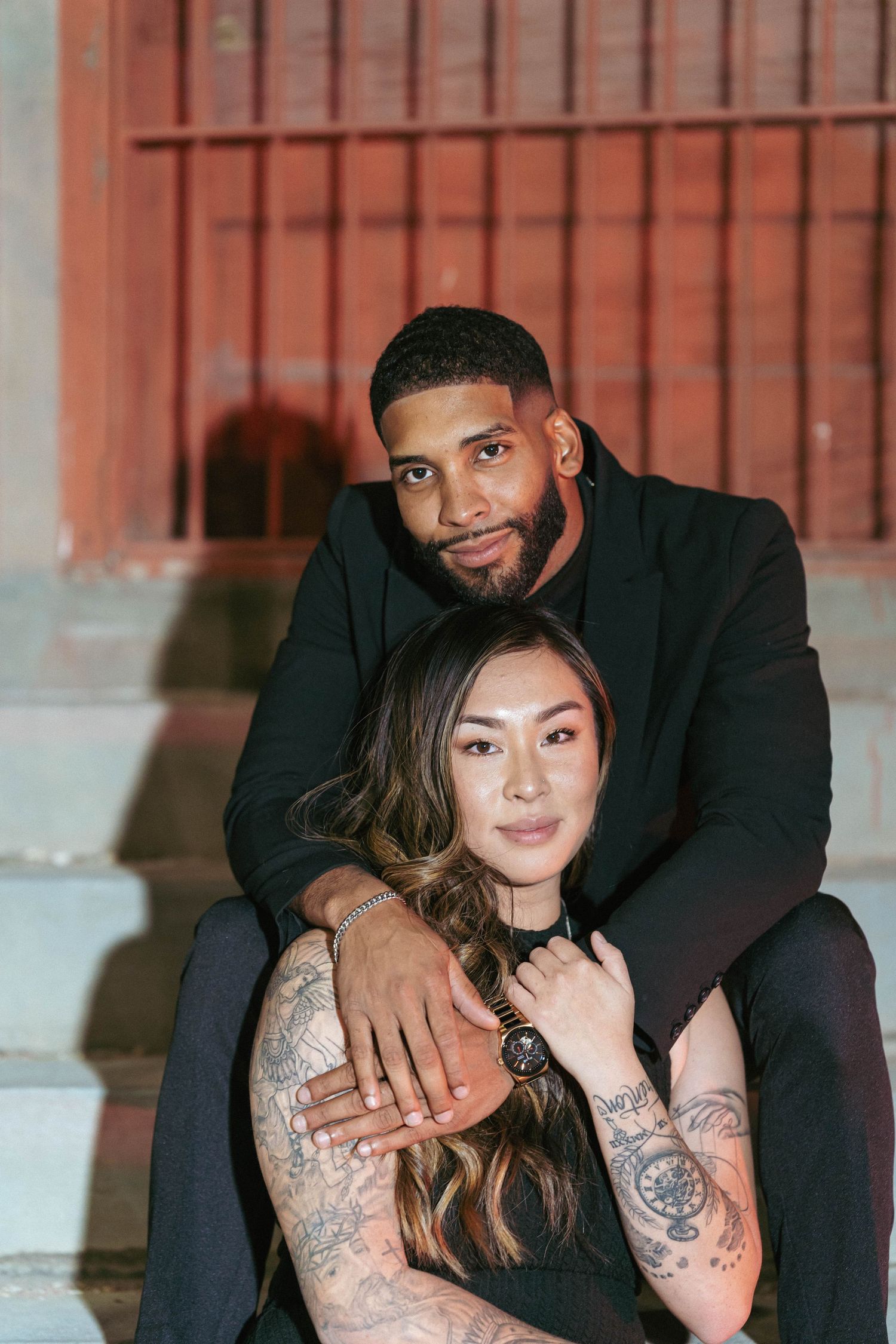 Intimate couple portrait session with woman in black dress and visible tattoos on brick background.
