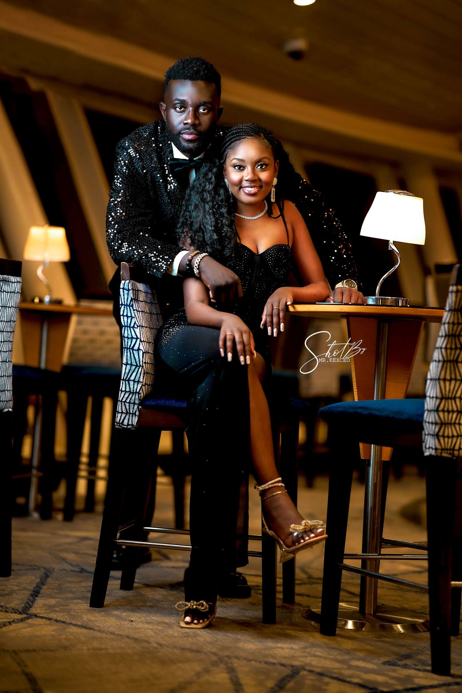 Stylish couple poses on floor during indoor photoshoot with ambient lighting.