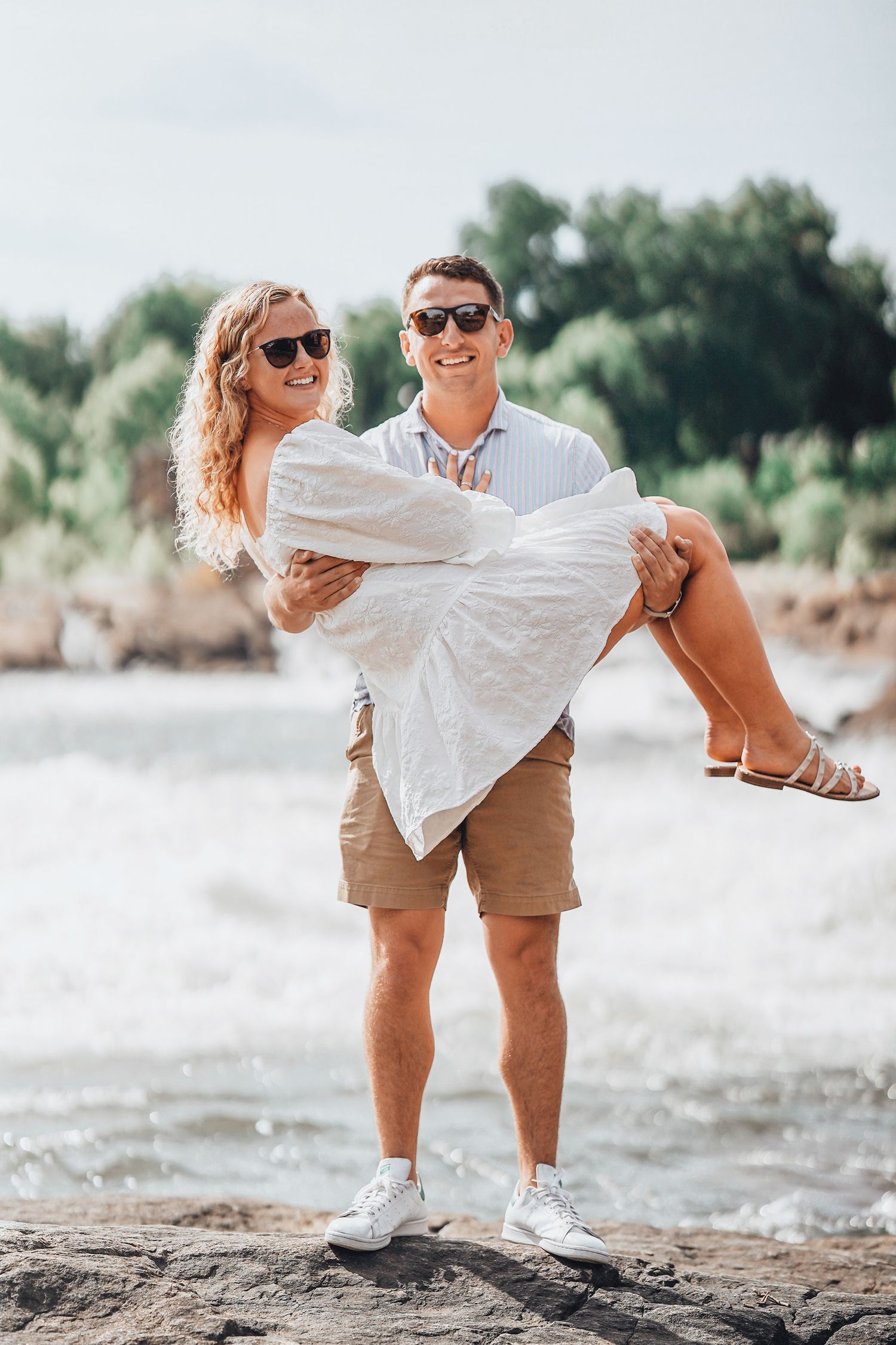 Man carrying woman in white dress on rocky beach during sunny day.