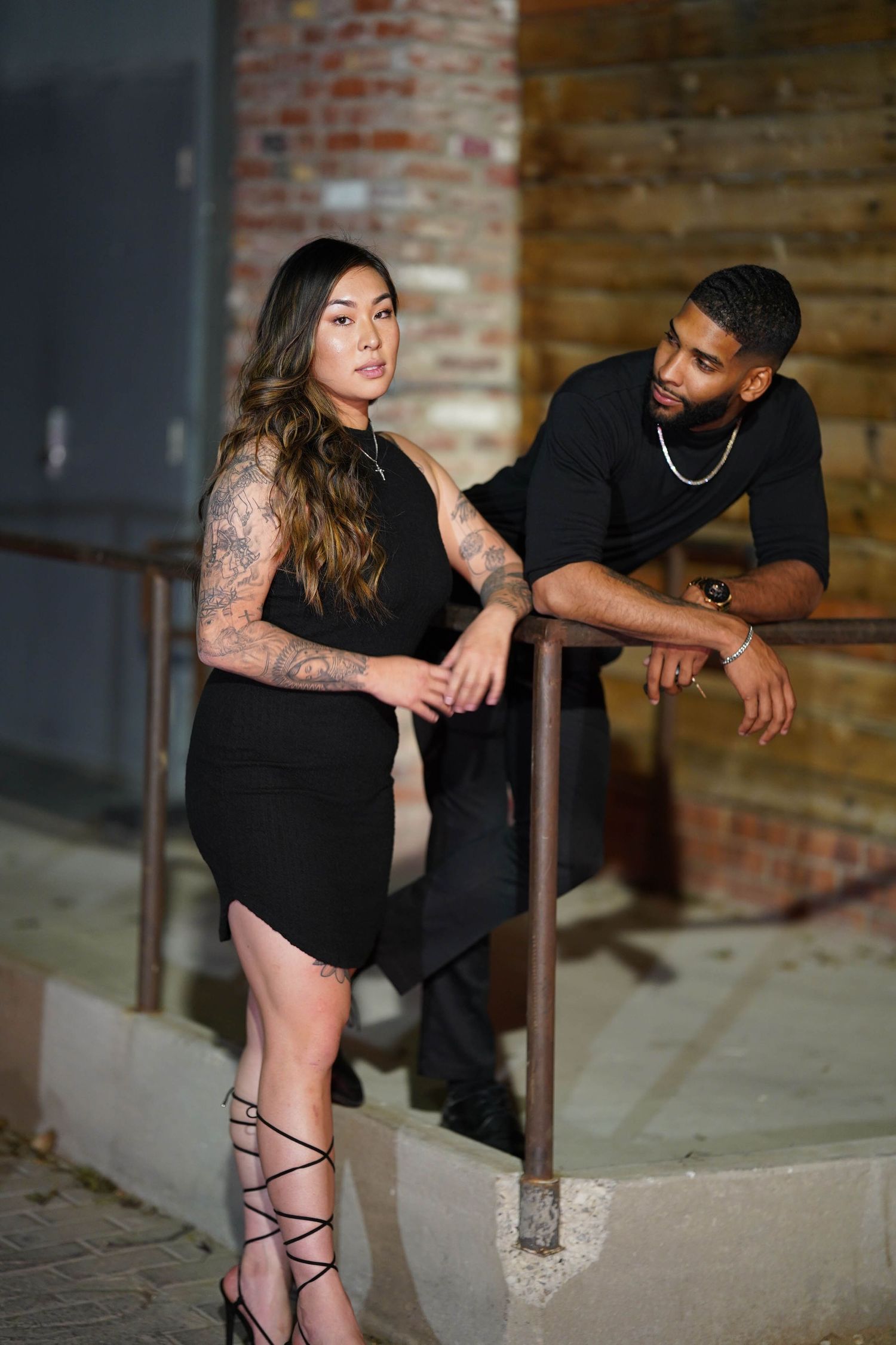Urban photoshoot of couple in all black against industrial brick backdrop.