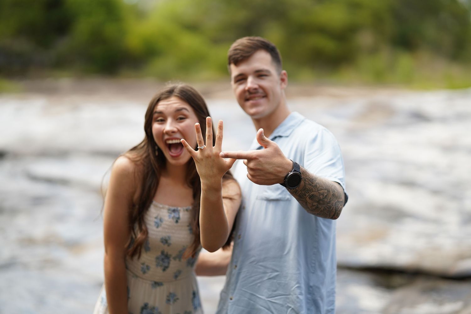 Two people excitedly showing off an engagement ring by a waterfall.