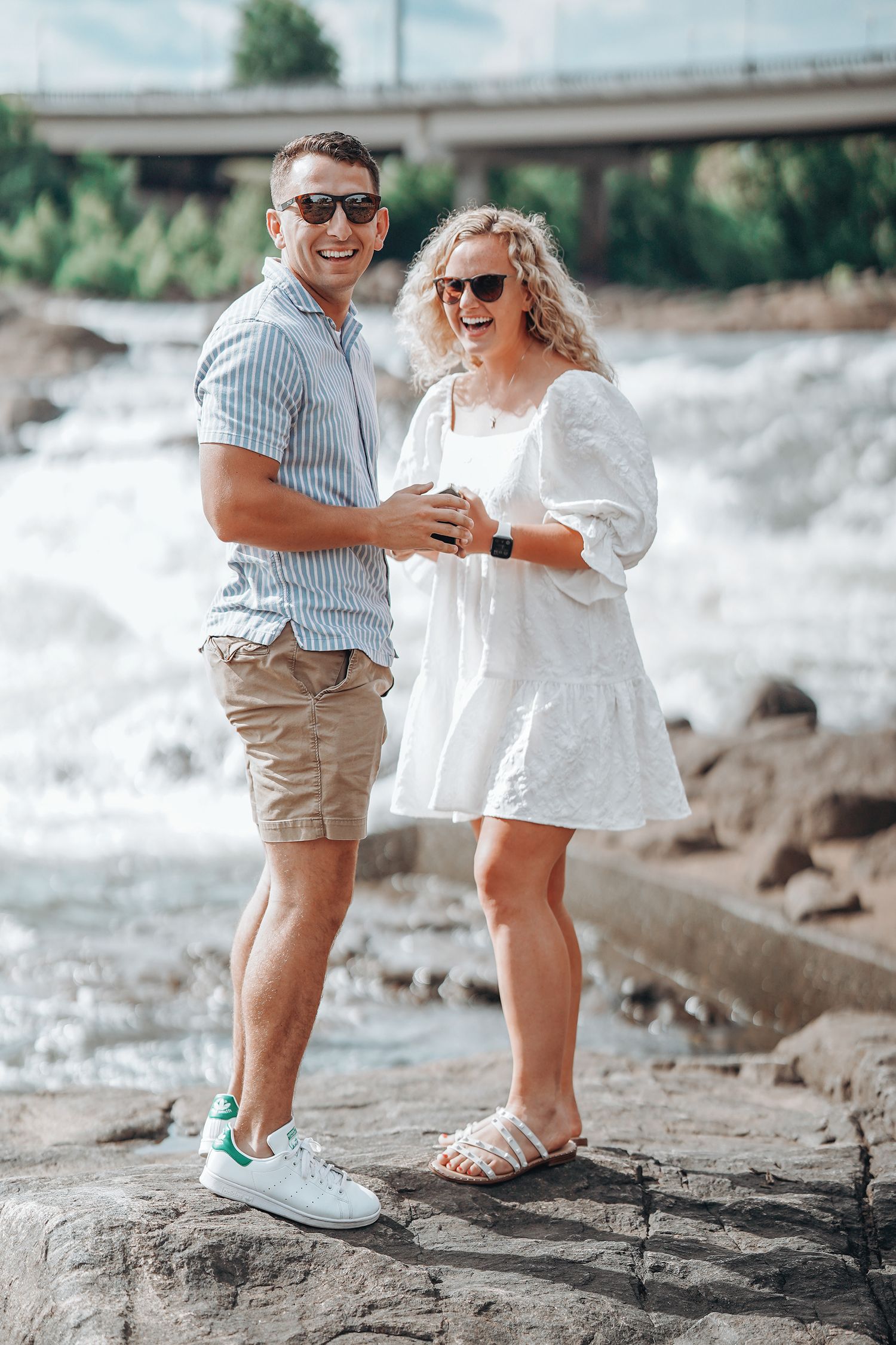 Couple in white outfits posing playfully on rocky waterfront.
