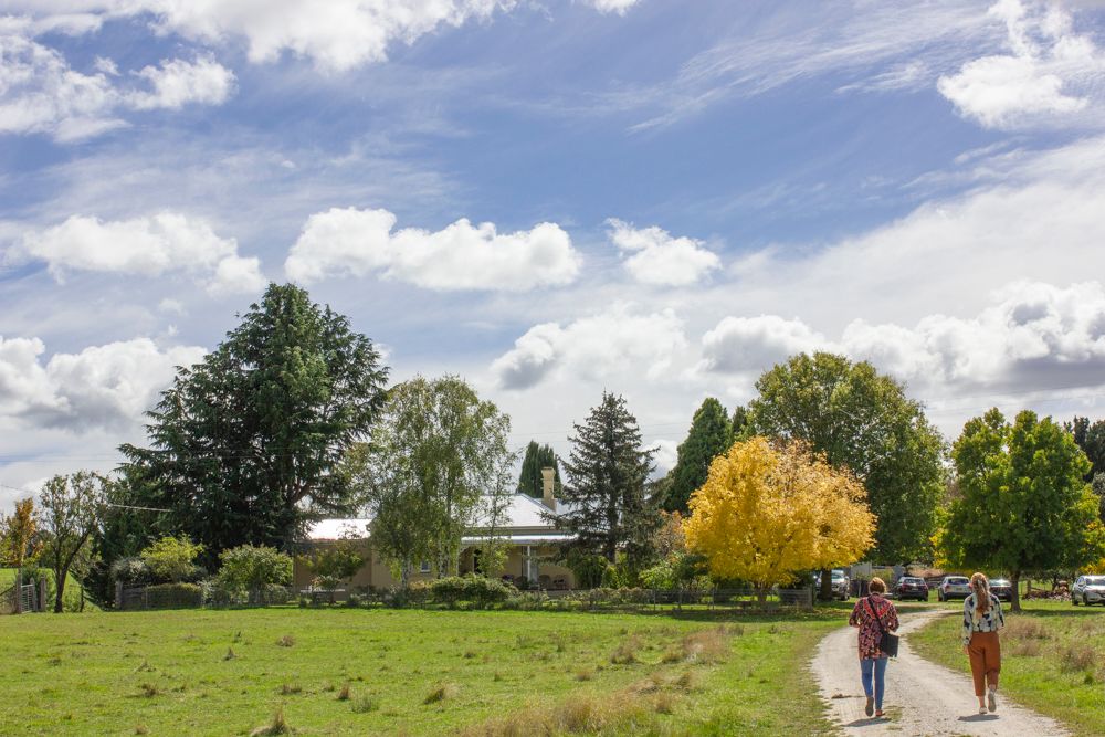 Two women walking on country dirt road toward historic farmhouse surrounded by autumn trees, green fields, blue sky with clouds.