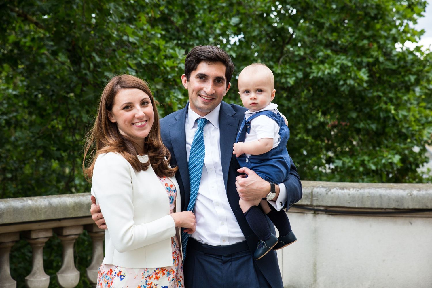 A family poses outdoors on a stone terrace with green trees in the background.