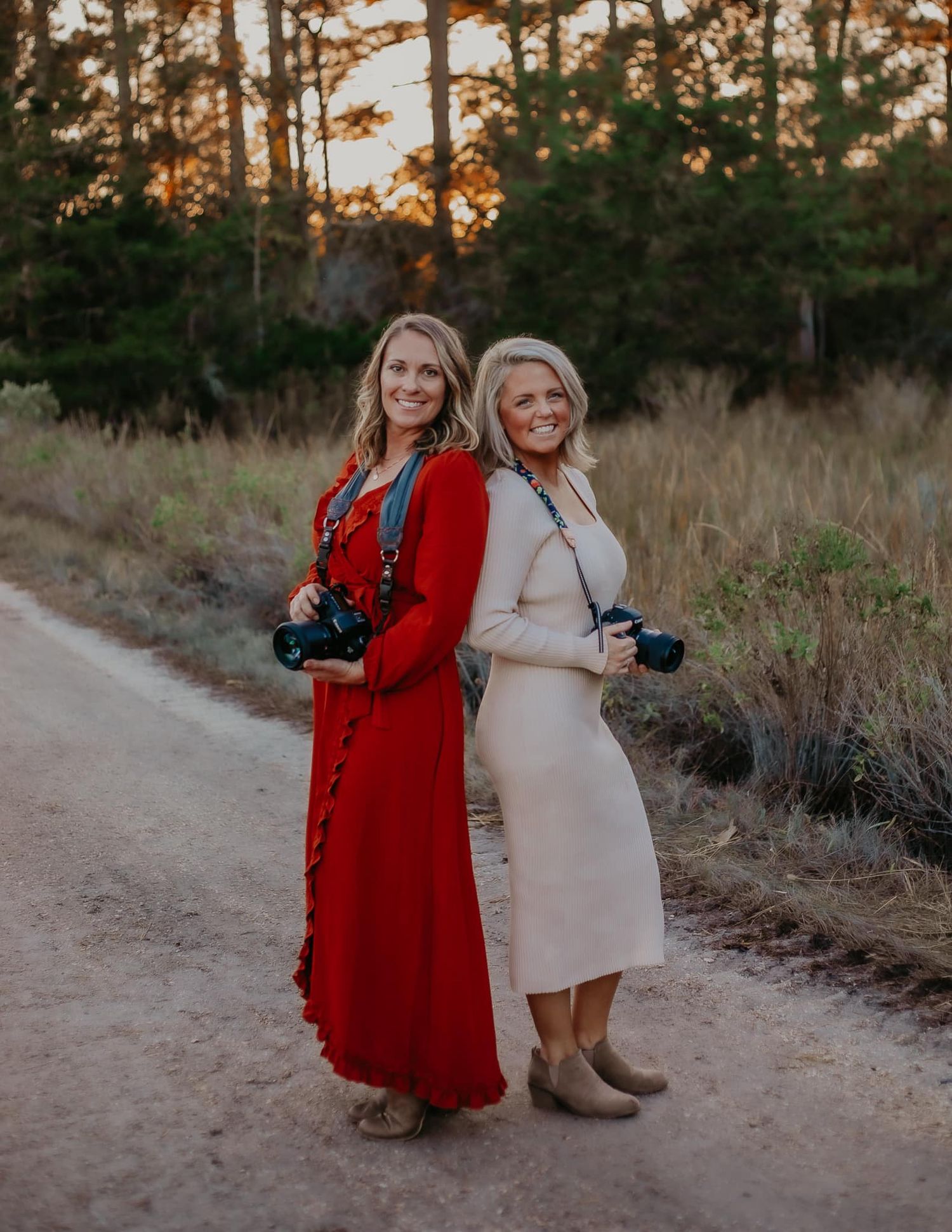 Two female photographers in red and cream dresses pose back-to-back on a dirt path at golden hour.